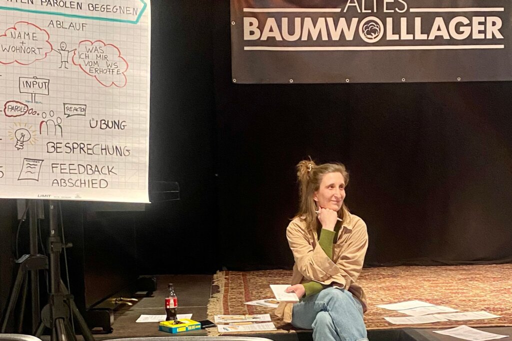 Moderatorin sitzt auf einer Bühne, um sie herum verstreut Zettel und Stifte und ein Getränk. Am linken Rand steht ein Flipchart mit einem skizzierten Trainings-Ablauf.