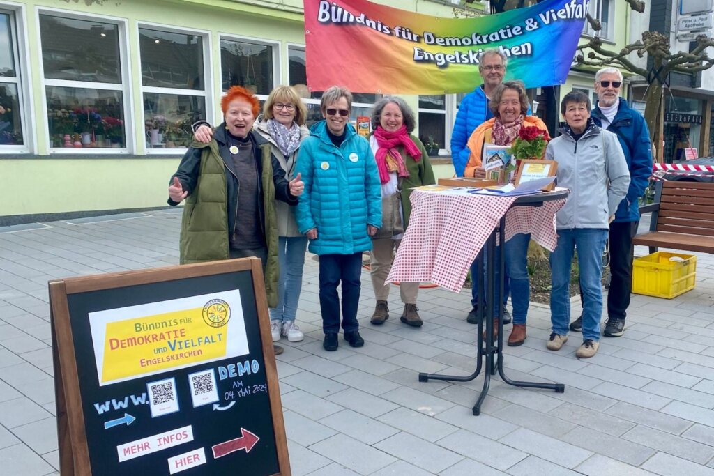 Eine Gruppe von 8 Menschen hinter einem Stehtisch auf einem Marktplatz. Im Hintergrund ein Banner mit der Aufschrift "Bündnis für Demokratie und Vielfalt"
Im Vordergrund steht eine Infotafel mit Informationen zur Demo am 4.Mai 2024